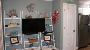 Una sala de estar con un televisor de pantalla plana sobre un mueble blanco, rodeada de estanterías con objetos decorativos y una guirnalda roja en la pared.