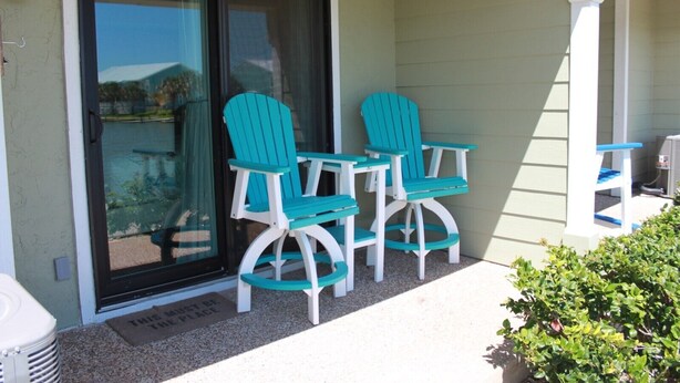 Un patio con dos sillas Adirondack color turquesa y una mesita, una puerta corrediza de vidrio, y una barandilla de porche blanca.
