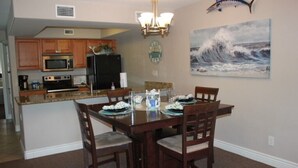 Un coin repas avec une table en bois, des chaises, un lustre, une cuisine en arrière-plan et un tableau représentant une vague accroché au mur.