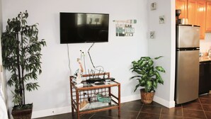 A living room with a flat-screen TV mounted on the wall, a bamboo plant, a wicker side table with electronic devices, and a kitchen area with wooden cabinets and a stainless steel refrigerator. - Corner condo with bay window overlooking pool (Rockport)