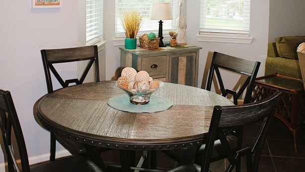 A dining area with a round wooden table, four chairs, a window with blinds, and wall decorations including a starfish painting and a name sign. - Corner condo with bay window overlooking pool (Rockport)