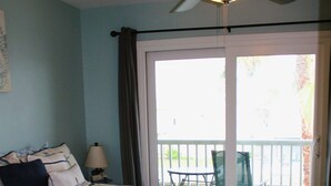 A bedroom with a bed, a ceiling fan, a nightstand, a lamp, and a view of a balcony with chairs. - Corner condo with bay window overlooking pool (Rockport)