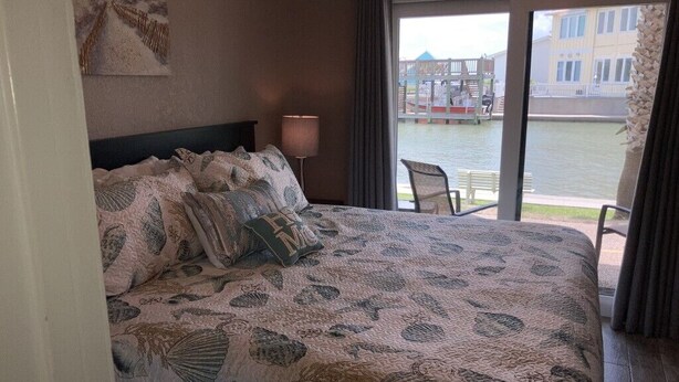A bedroom with a large bed, a view of a water body, and a chair by the window. - Beautiful Ground Level Condo (Rockport)
