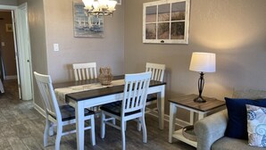 A dining area with a wooden table and white chairs, a chandelier, and a painting of a beach scene. - Beautiful Ground Level Condo (Rockport)