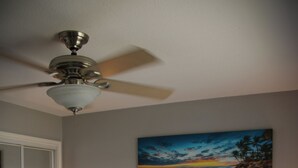 Een slaapkamer met een bed, een plafondventilator, een schilderij van een strandlandschap en een nachtkastje met een lamp.