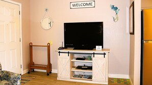 Een kamer met een televisie op een houten standaard, een 'WELCOME' bord, een spiegel aan de muur en een lijst met een afbeelding.