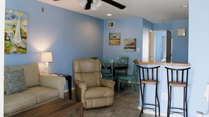 Un salon comprenant un canapé, un fauteuil inclinable, une table à manger avec des chaises, un coin bar avec des tabourets, un ventilateur de plafond et des œuvres d’art accrochées aux murs.