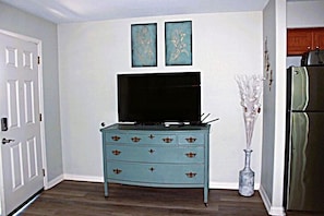 Una habitación con un aparador azul, un televisor de pantalla plana, un florero con ramas y un refrigerador.