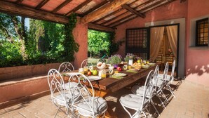 Un área de comedor al aire libre con una larga mesa de madera, sillas de metal blanco y una pared de ladrillo.