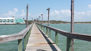 Un muelle de madera se adentra en aguas tranquilas, con un edificio a la izquierda y un cielo despejado.