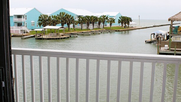A balcony with a view of a water body, a dock with a boat, and a building with a blue exterior. - Second floor condo with canal view (Rockport)