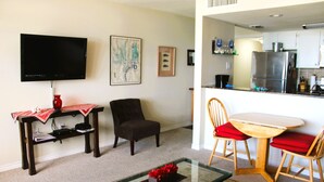 Una sala de estar con televisor, una mesa de comedor con sillas, una mesa de centro y una cocina al fondo.