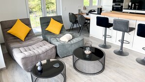 A modern living room with a grey sofa, ottoman, and round tables, a kitchen with white cabinets and a black countertop, and a balcony with a railing. - Gemütliche & Luxuriöse Ferienwohnung in Top-lage (Höchenschwand)