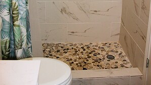 A shower with a rain showerhead, a pebble-patterned floor, and a shower curtain with a tropical leaf design. - Fantastic water view from comfortable cozy condo (Rockport)