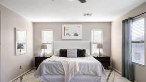 A bedroom with a large bed, two bedside tables, a ceiling fan, and a window with blinds. - "THE HAMPTON" Gilbert I Designer Style + Comfort I 3bed/2.5bath (Gilbert)