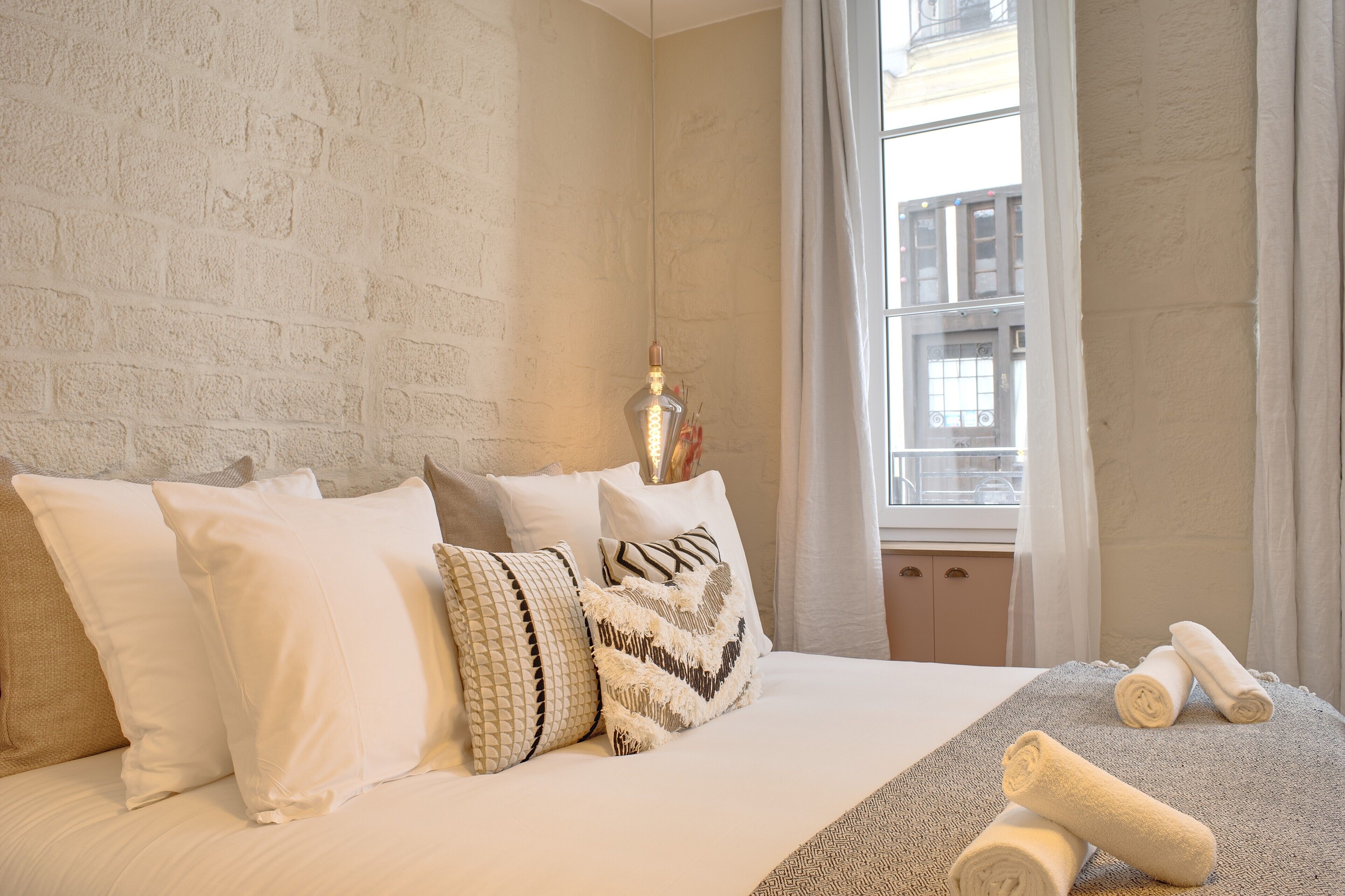 A neatly made bed with white bedding and patterned pillows, a window with curtains, and a view of buildings outside.