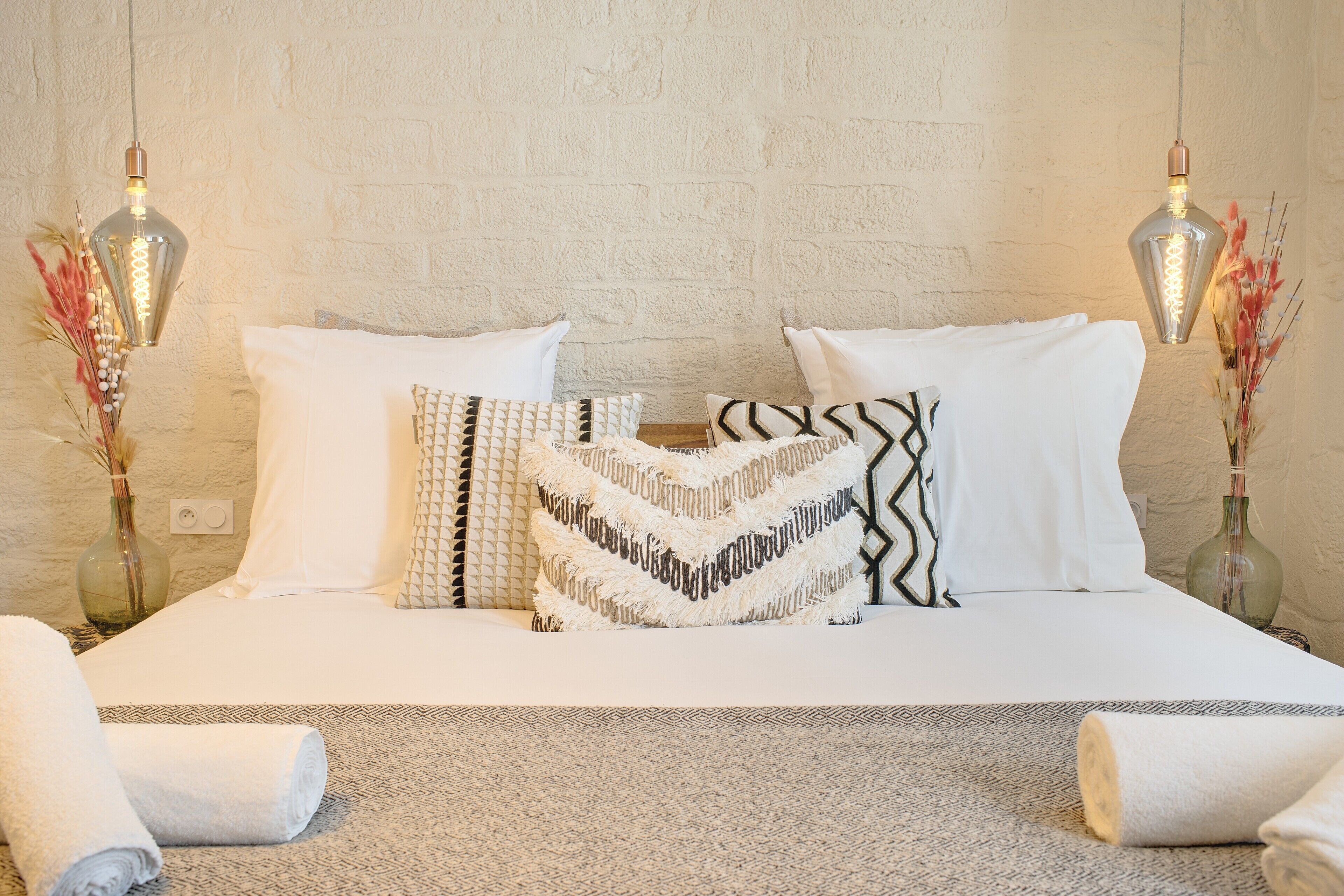 Una cama bien tendida con sábanas blancas y almohadas estampadas, una alfombra con textura y dos lámparas de pared con jarrones decorativos.
