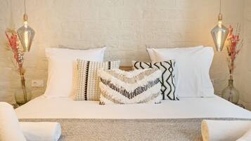 Una cama bien tendida con sábanas blancas y almohadas estampadas, una alfombra con textura y dos lámparas de pared con jarrones decorativos.