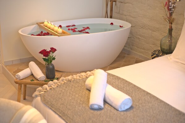 Un espace spa comprenant une grande baignoire remplie d’eau et de pétales de rose, un plateau en bois avec des bougies et des serviettes enroulées posées sur une table.