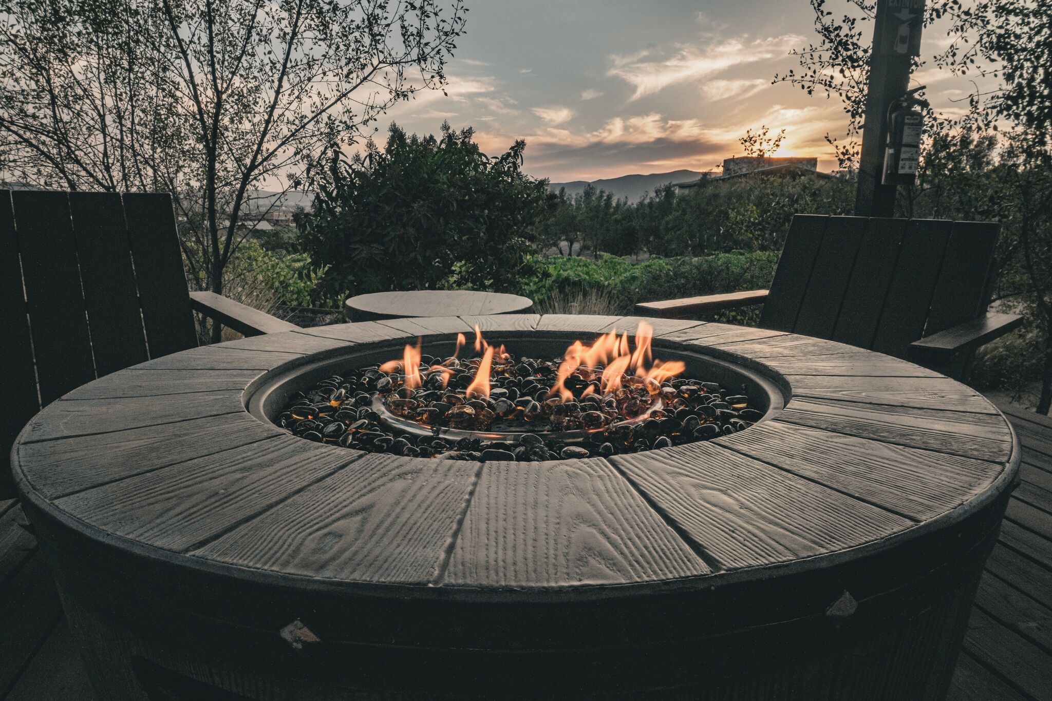 Una chimenea de madera circular con llamas, rodeada de mobiliario de exterior en una terraza.