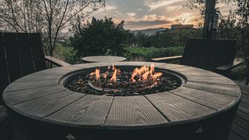 Una chimenea de madera circular con llamas, rodeada de mobiliario de exterior en una terraza.