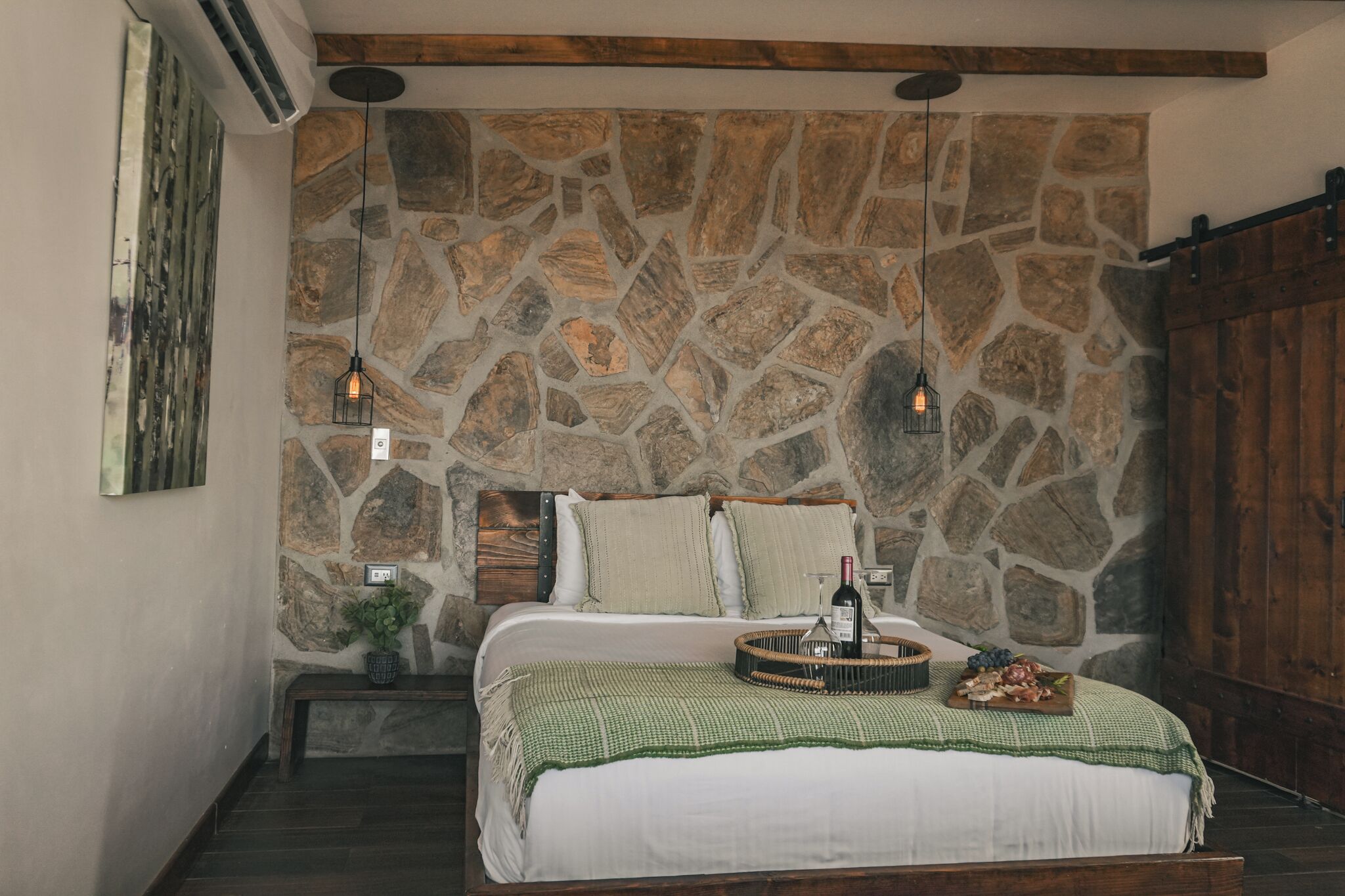 A bedroom with a stone wall, a bed with a green checkered blanket, a bedside table with a bottle, and a basket of fruit.