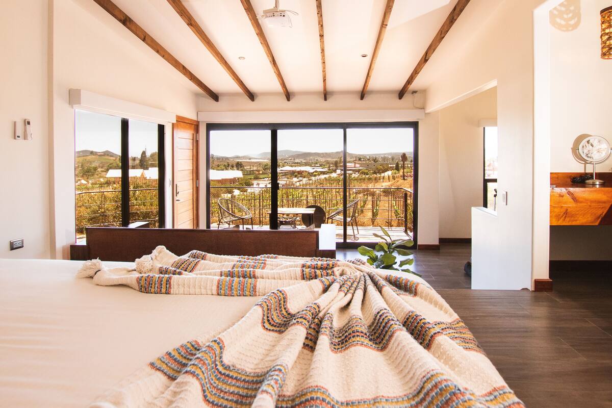 Une chambre spacieuse avec un grand lit, une vue sur les vignes grâce à de grandes fenêtres du sol au plafond et un plafond en bois.