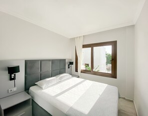 A modern hotel room with a large bed, a nightstand, a wall-mounted lamp, and a window with a view of buildings and trees. - Turgutreis Apart Otel (Bodrum)
