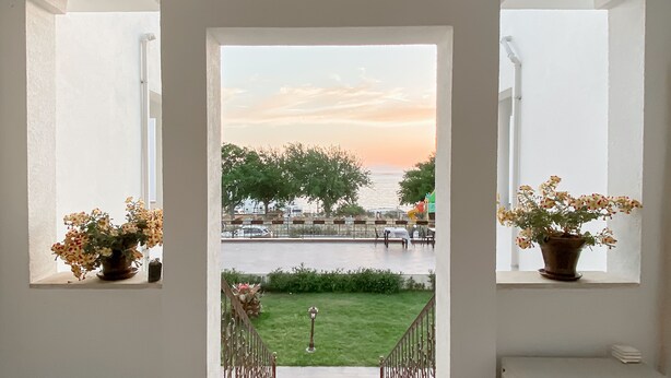 A view through a doorway with two windows, each with a potted plant. There is a grassy area in front of the windows, and a body of water with trees and buildings in the background. - Turgutreis Apart Otel (Bodrum)