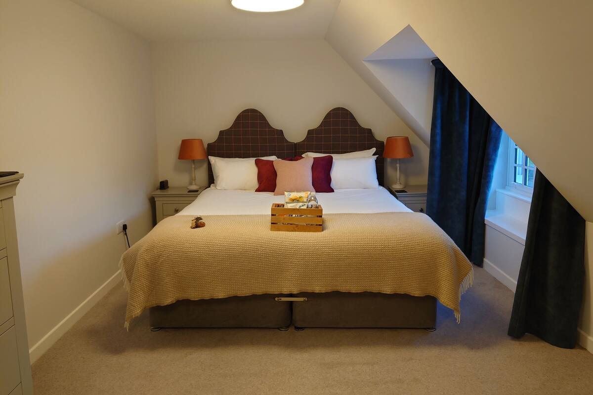 A neatly made bed with a headboard, two bedside tables with lamps, a small wooden box on the bed, and a window with curtains.