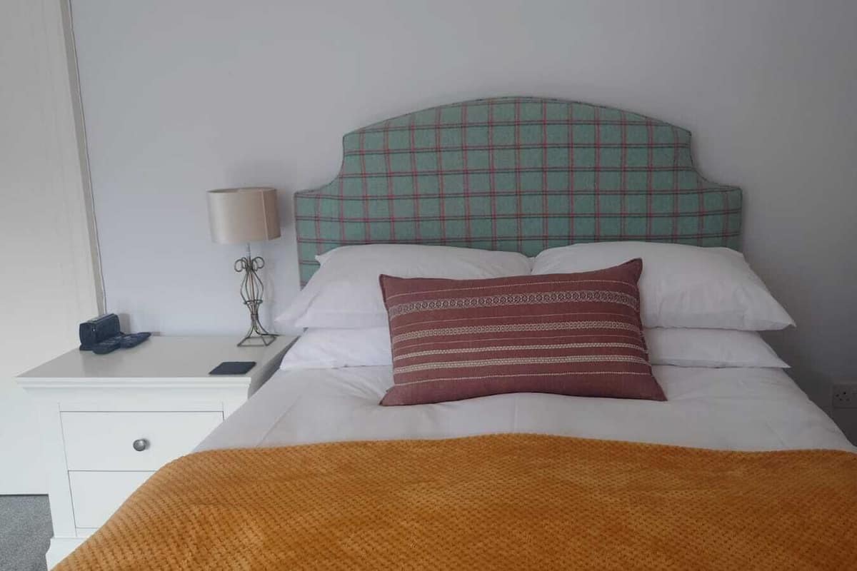 A neatly made bed with a plaid headboard, a bedside lamp, and a small nightstand with a lamp and a phone.