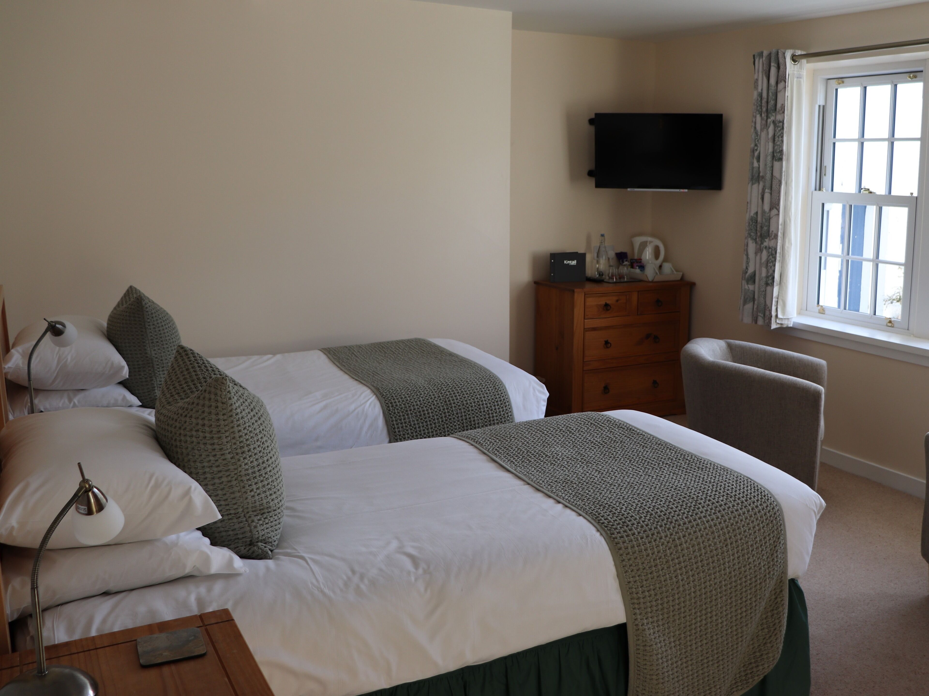 Een hotelkamer met twee bedden, een houten dressoir, een aan de muur bevestigde televisie en een raam met gordijnen.