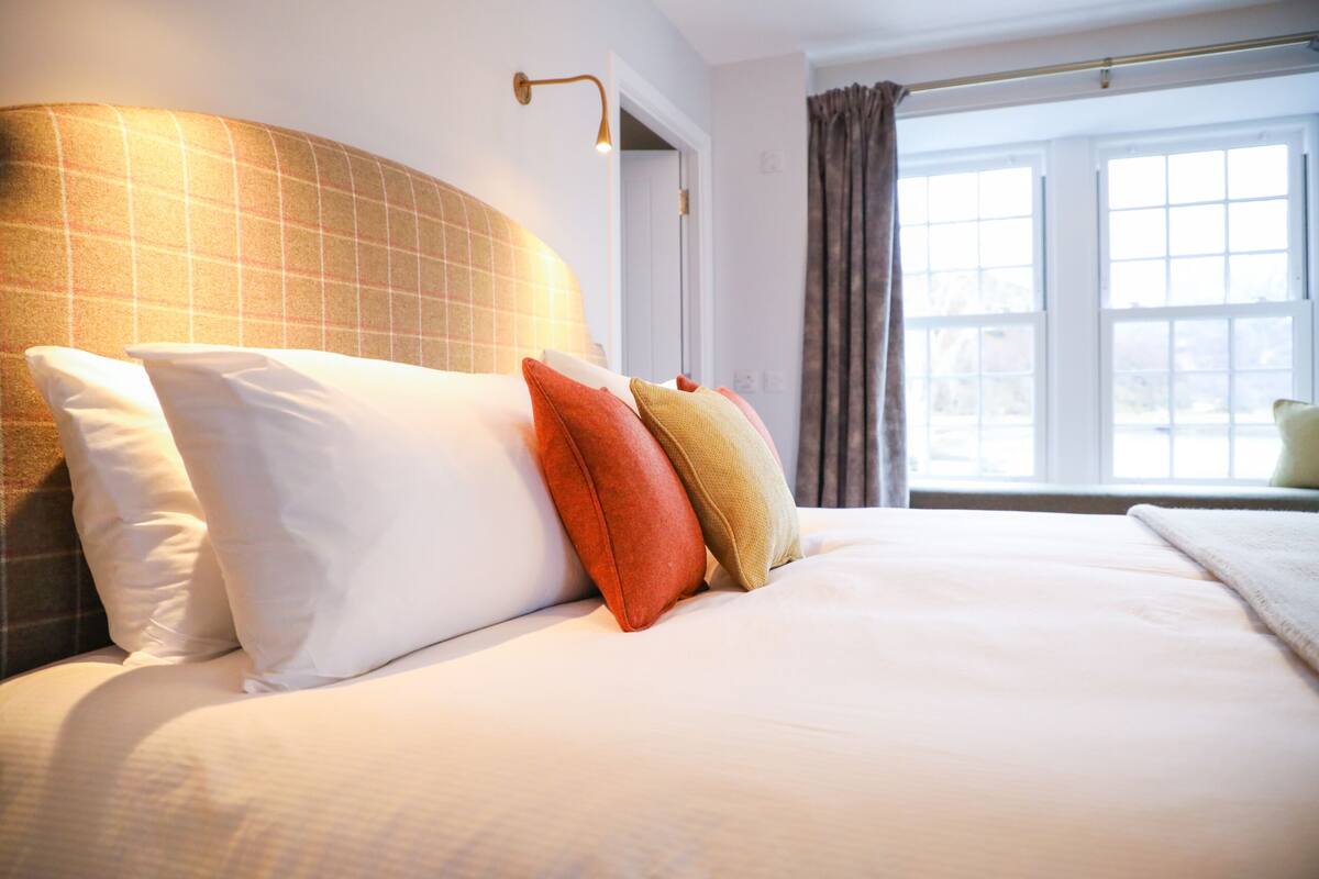A neatly made bed with white and colored pillows, a headboard with a checkered pattern, and a window with curtains.