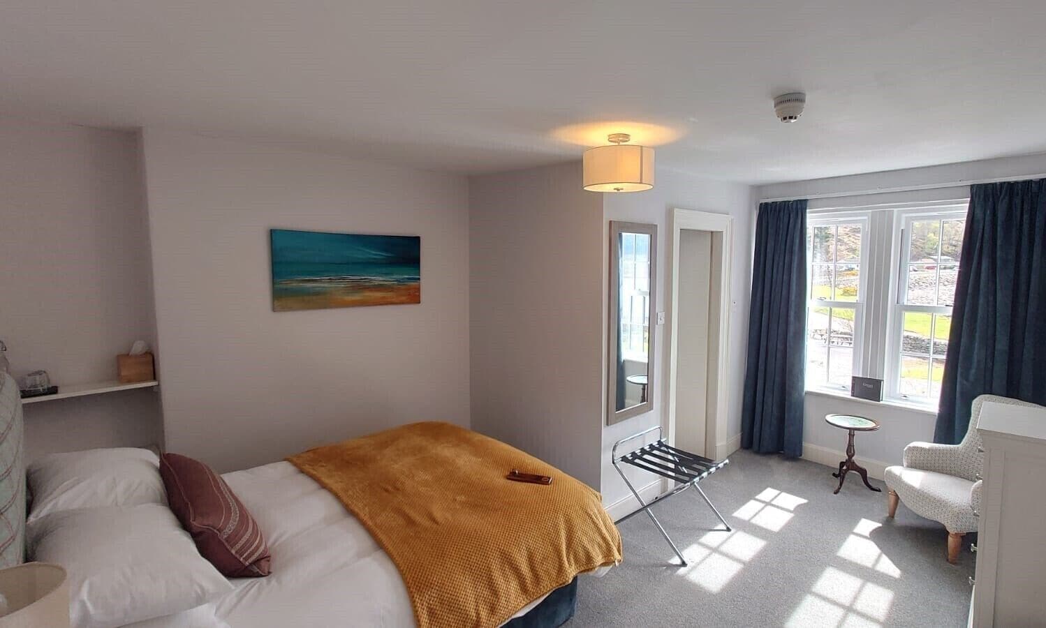 Una camera d'albergo con un letto, un tavolino, una sedia, uno stendibiancheria, un quadro e una finestra con tende.