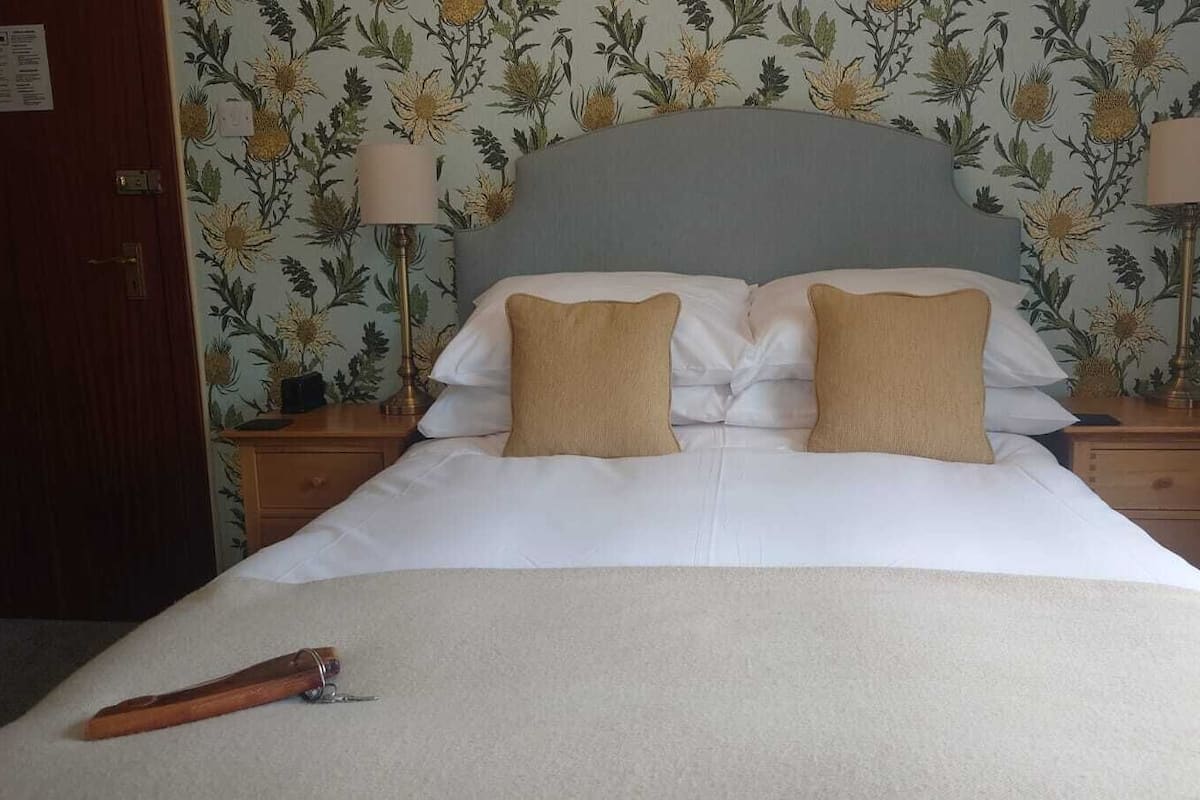 A neatly made bed with two pillows, a wooden nightstand with a lamp, and floral wallpaper.