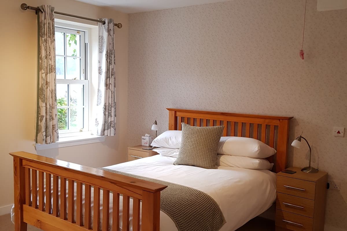 A bedroom with a wooden bed, a nightstand with a lamp, a window with curtains, and a ceiling light fixture.