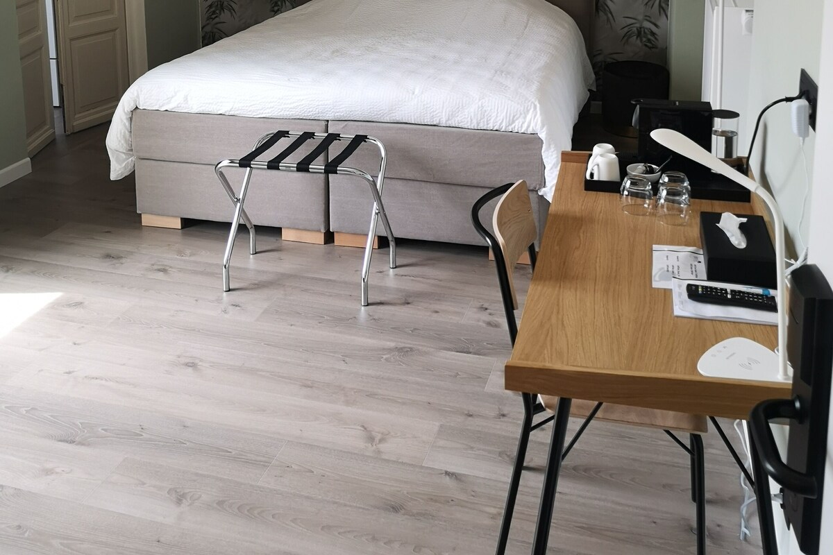 Un dormitorio con cama, escritorio con silla, lámpara y una mesita con vasos y un control remoto.