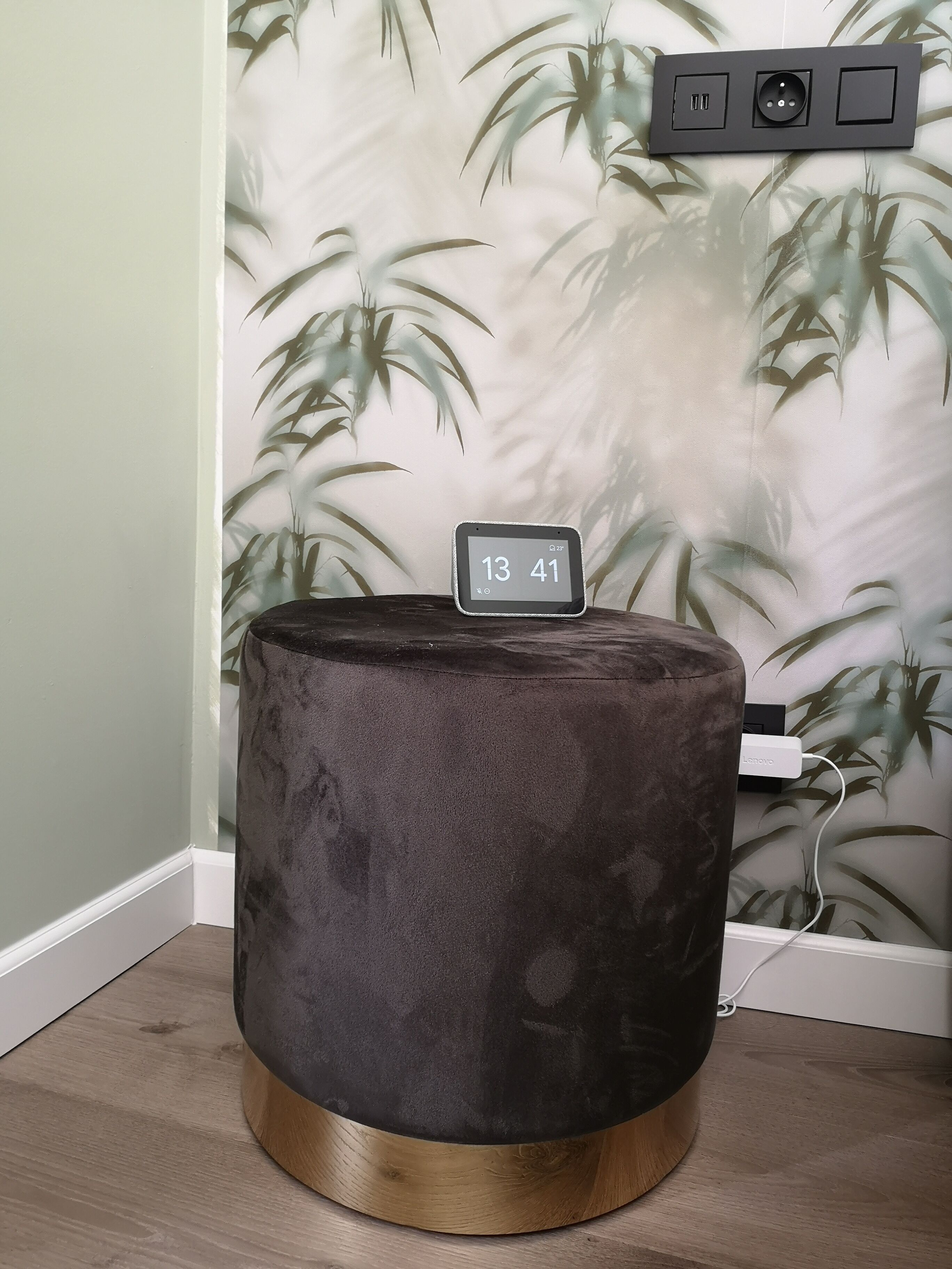 Un pouf rotondo e scuro con base in legno, posizionato in un angolo accanto a una parete con carta da parati a foglie di bambù.