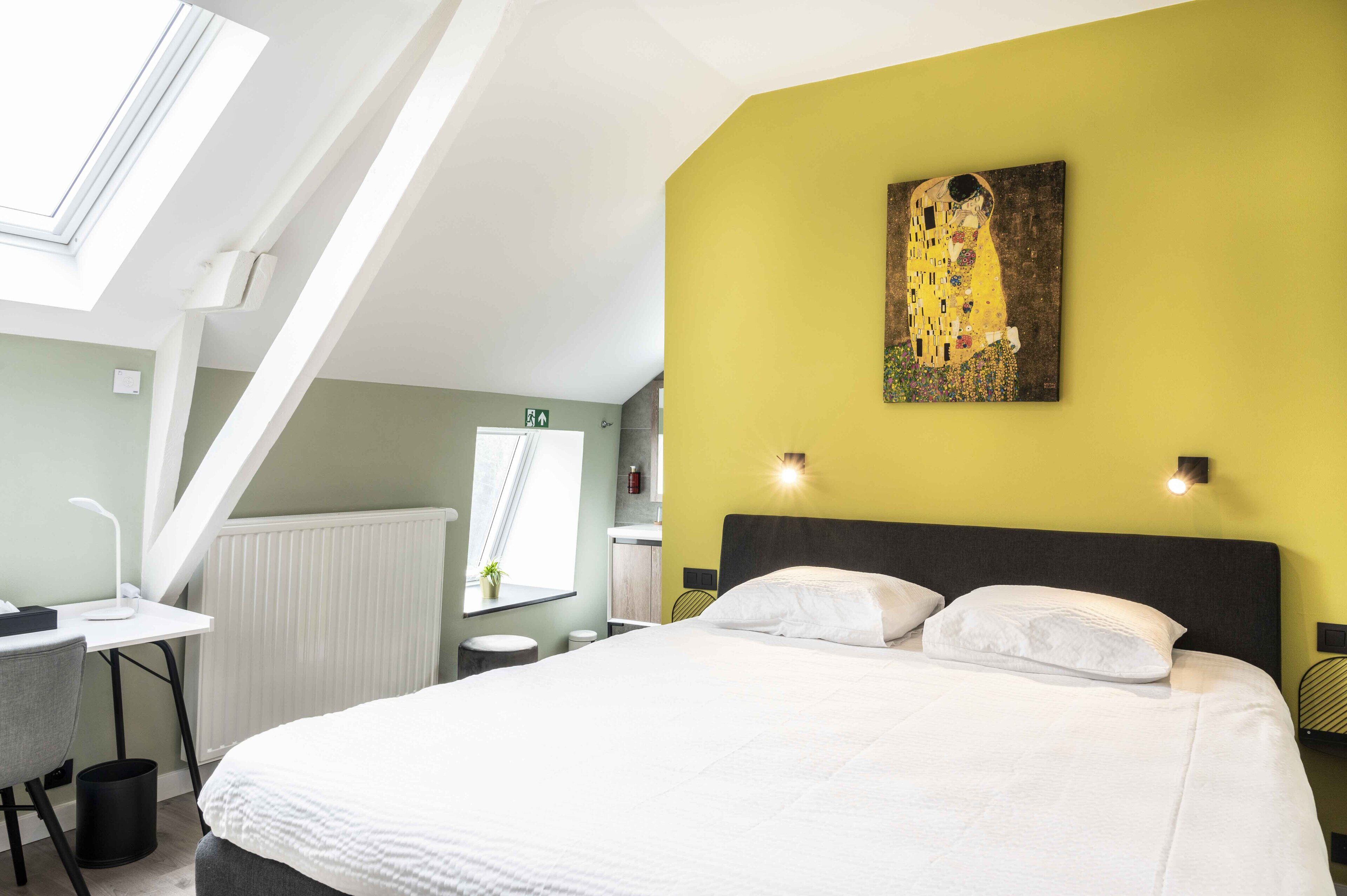 A modern hotel room with a large bed, a desk with a chair, a skylight, and a painting on the wall.