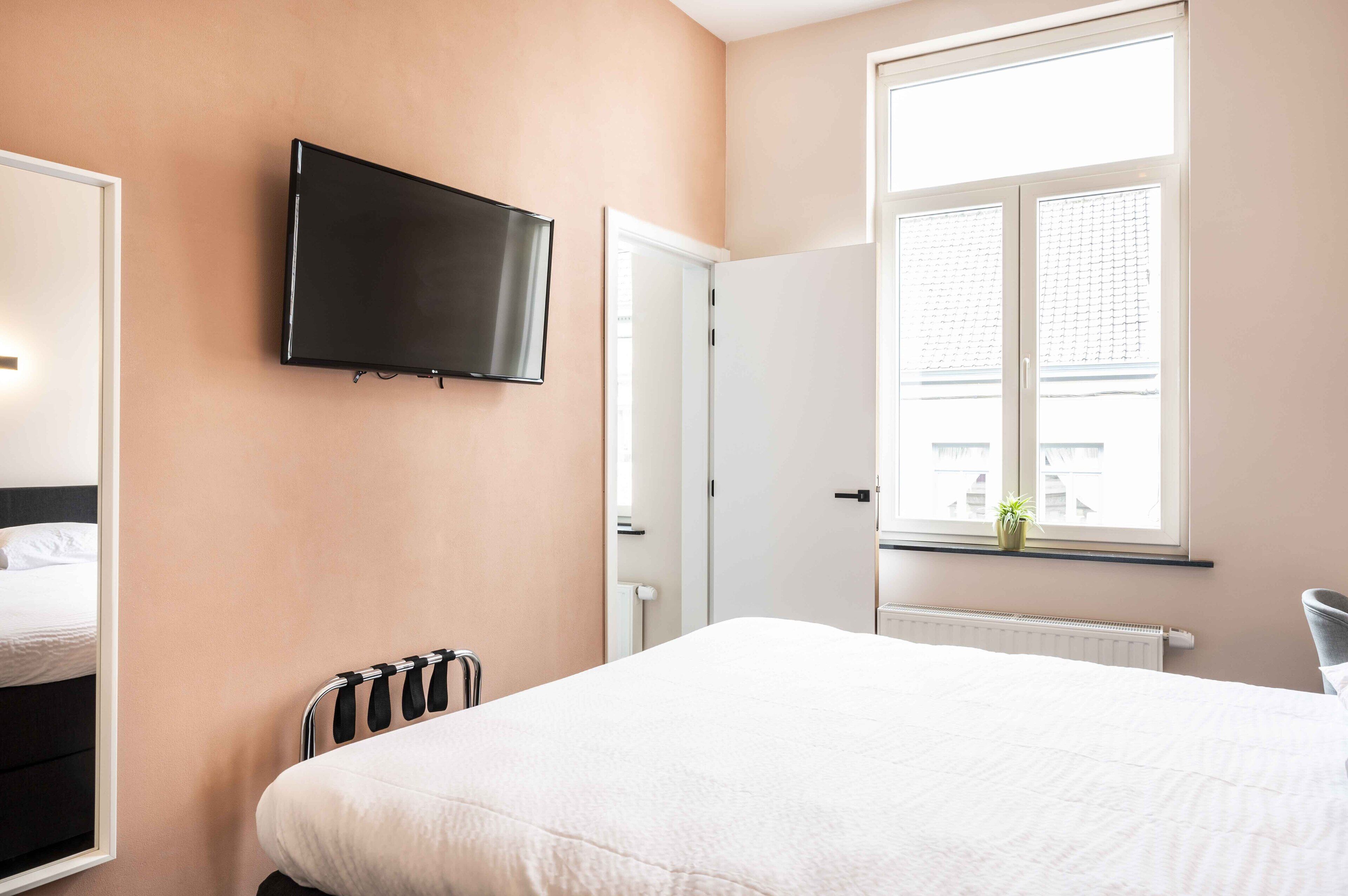 A hotel room with a bed, a TV mounted on the wall, a window with a view of buildings, and a radiator.
