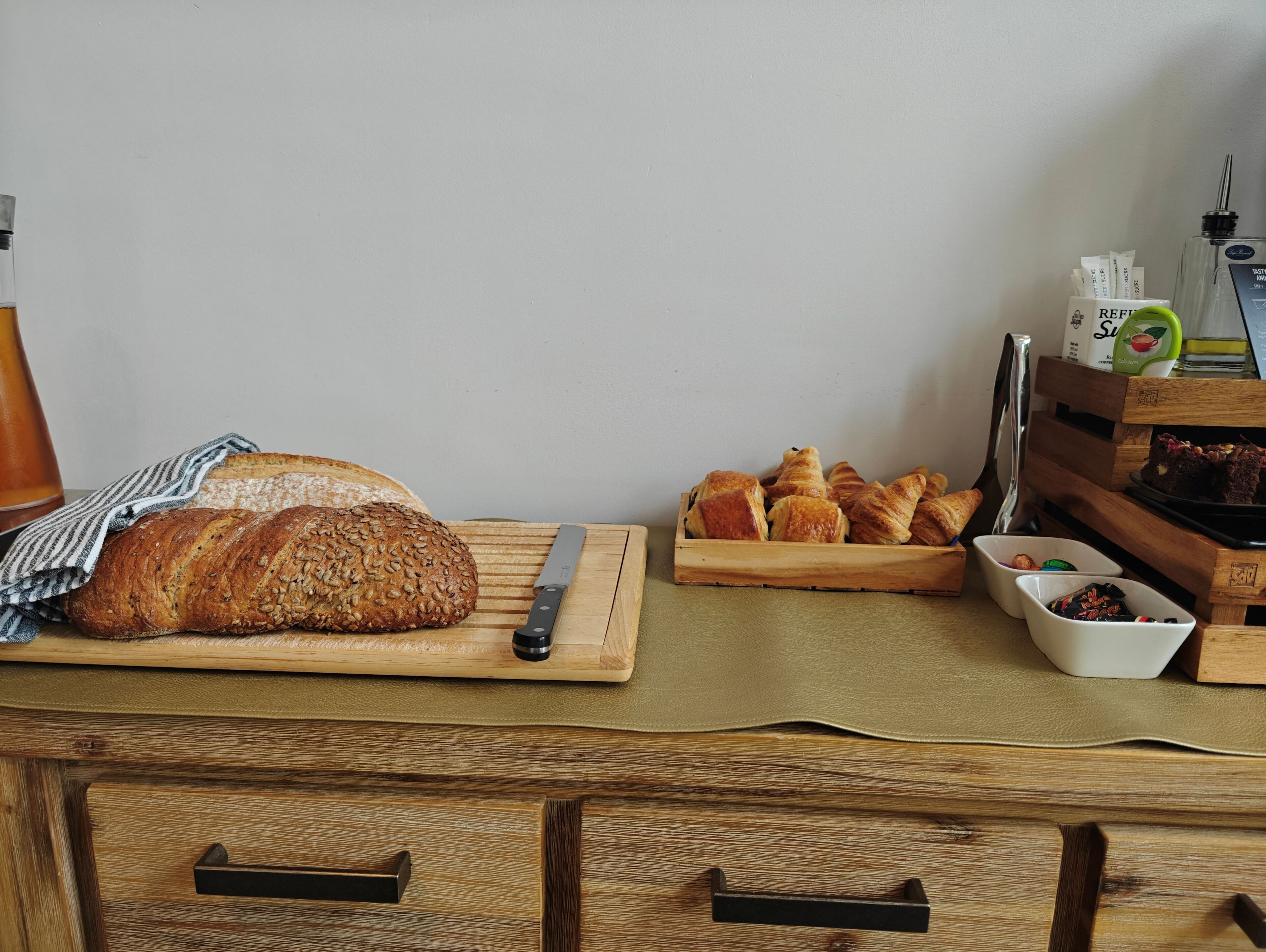 A wooden cutting board with a loaf of bread and a knife, a wooden tray with croissants, and a small bowl of nuts.