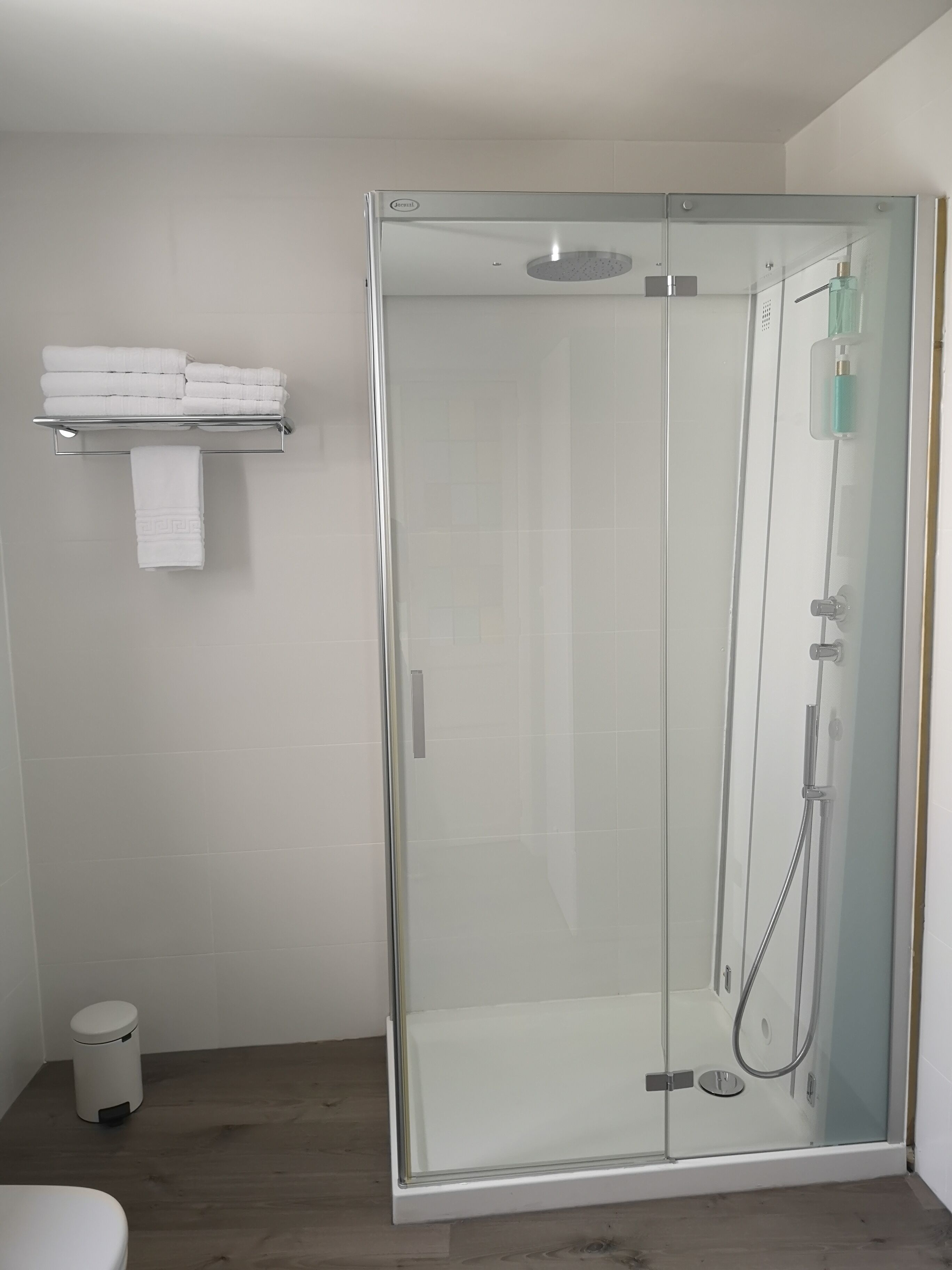 Een moderne badkamer met een douchecabine van glas, een betegelde muur in het wit, een klein plankje met opgevouwen handdoeken en een witte prullenbak.