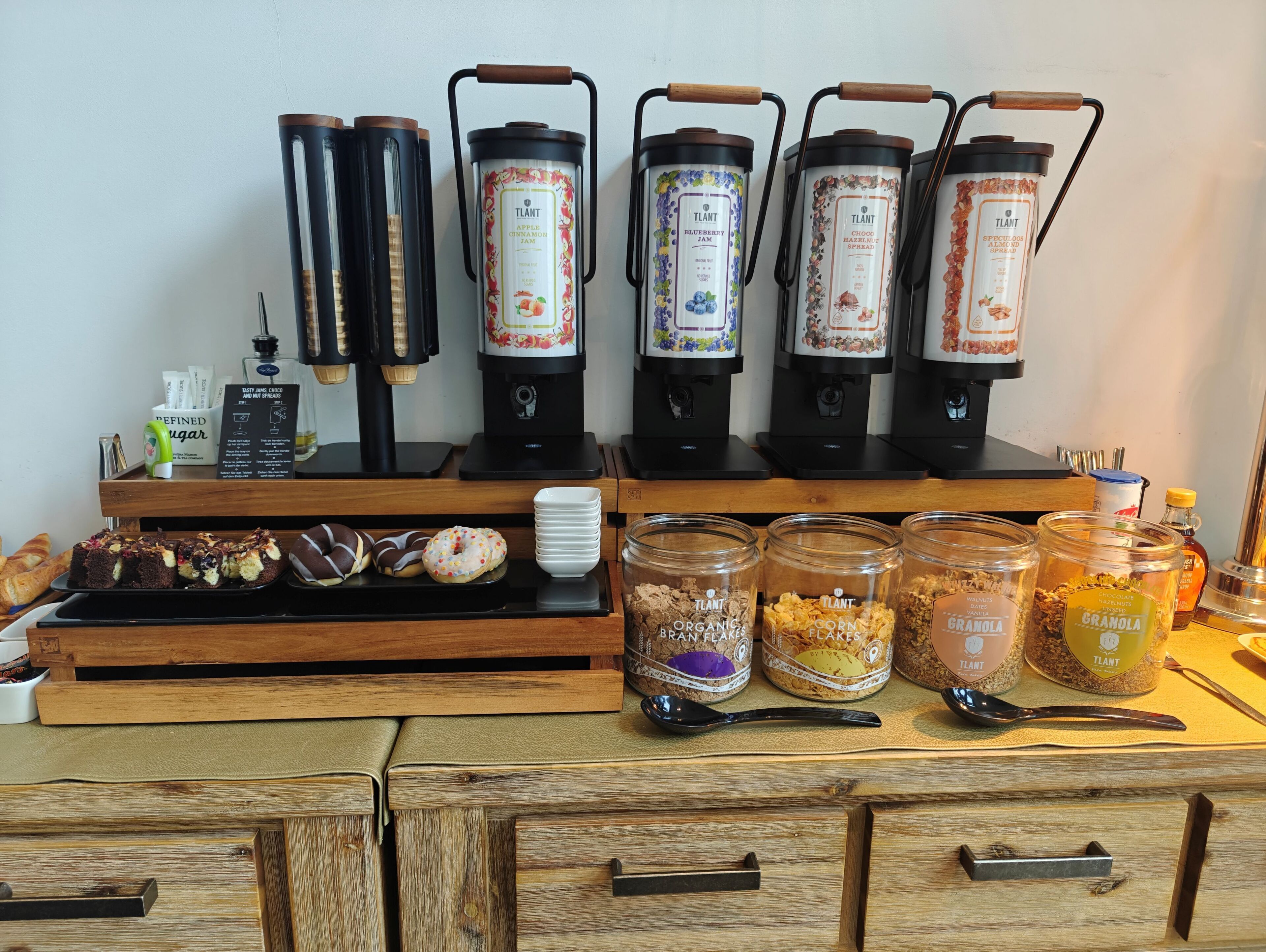 Una estación de café con varios dispensadores, una bandeja de pasteles y frascos con granola y otros bocadillos.