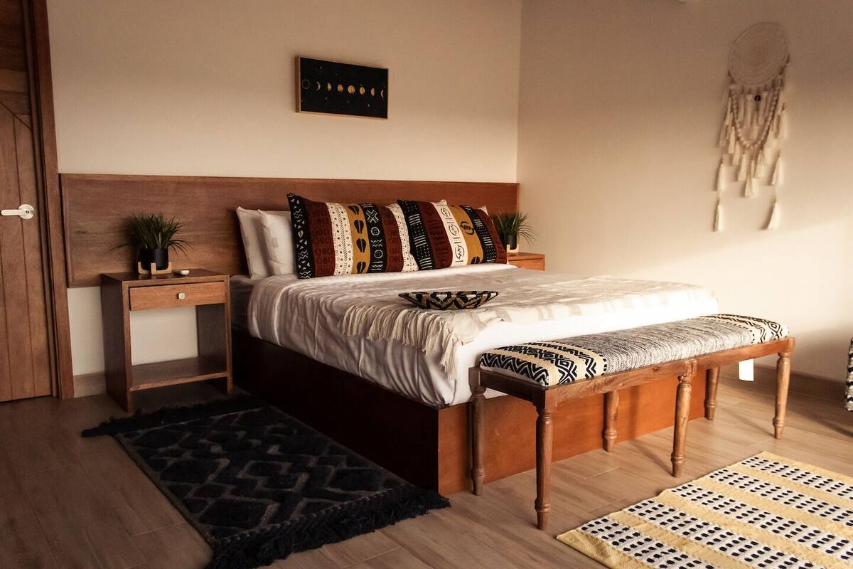 A bedroom with a wooden bed, a bedside table with a plant, a wall-mounted dreamcatcher, and a patterned rug.