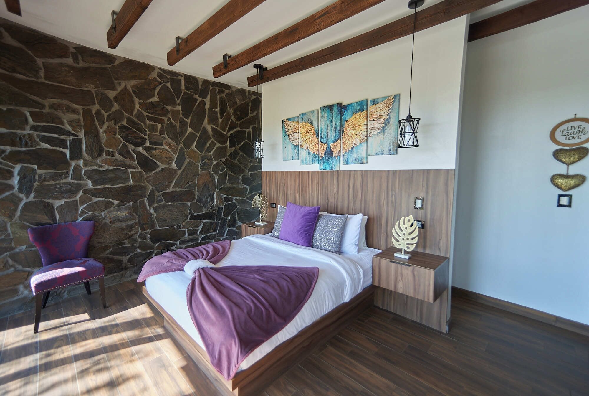 Un dormitorio con pared de piedra, una cama con manta púrpura, una mesita de noche y una silla.