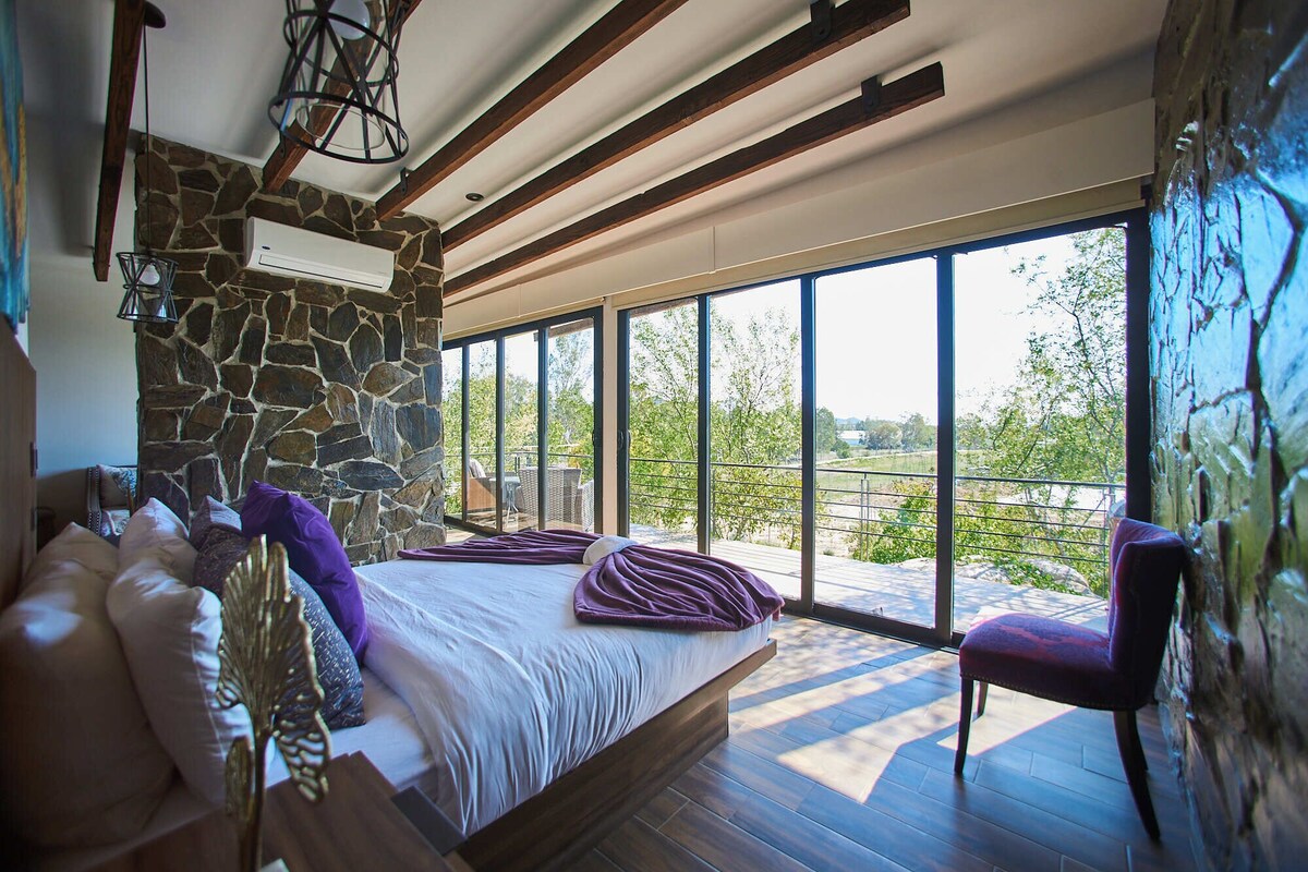 A bedroom with a stone wall, a bed with a purple blanket, a chair, and a view of the outdoors.
