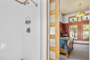A bathroom with white tiled walls, a shower, and a toilet. There is a wooden-framed window with a view of trees outside.