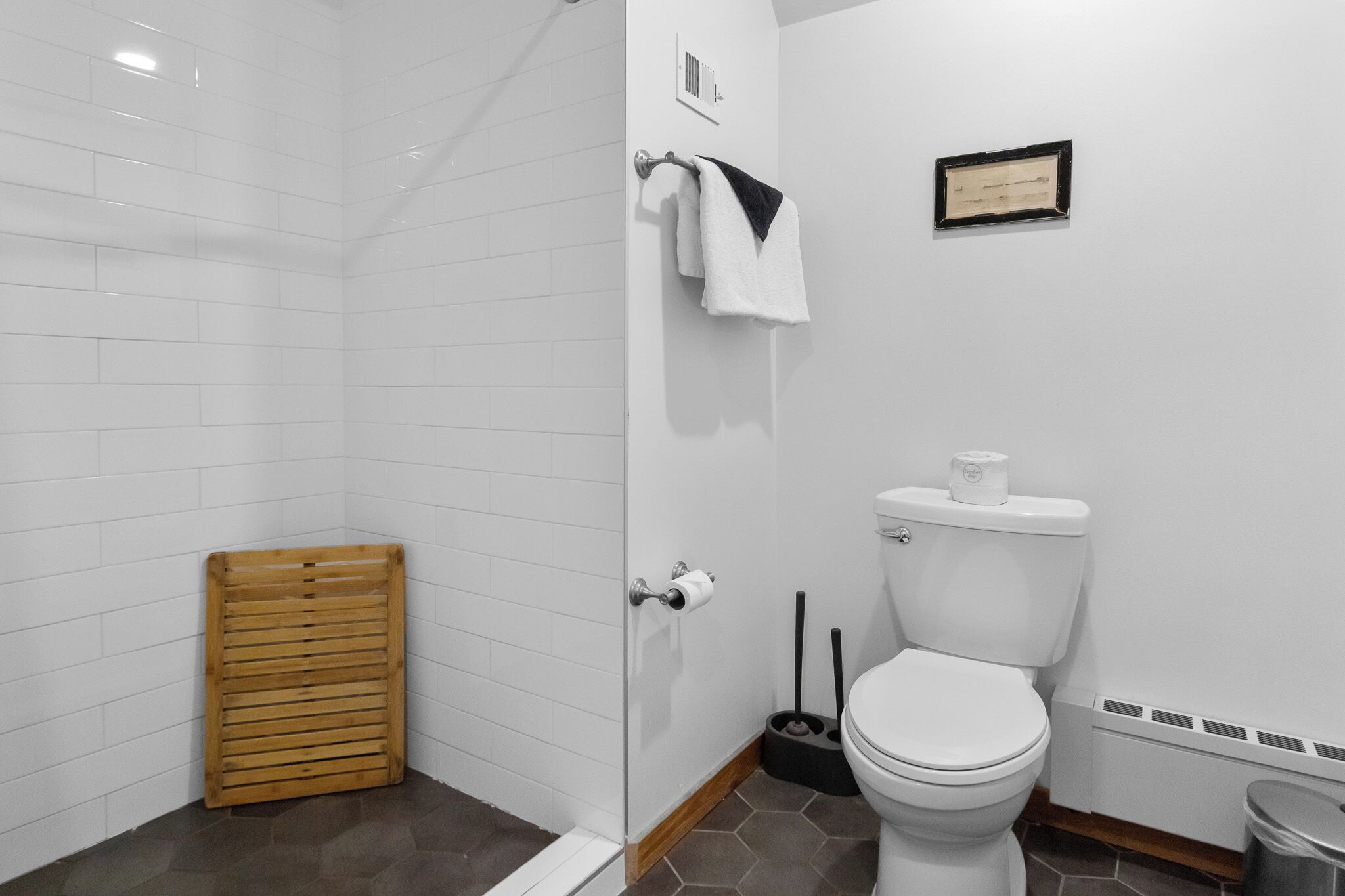 Un baño con ducha revestida de azulejos blancos, un banco de ducha de madera, un inodoro con un cepillo de limpieza negro y un certificado enmarcado en la pared.