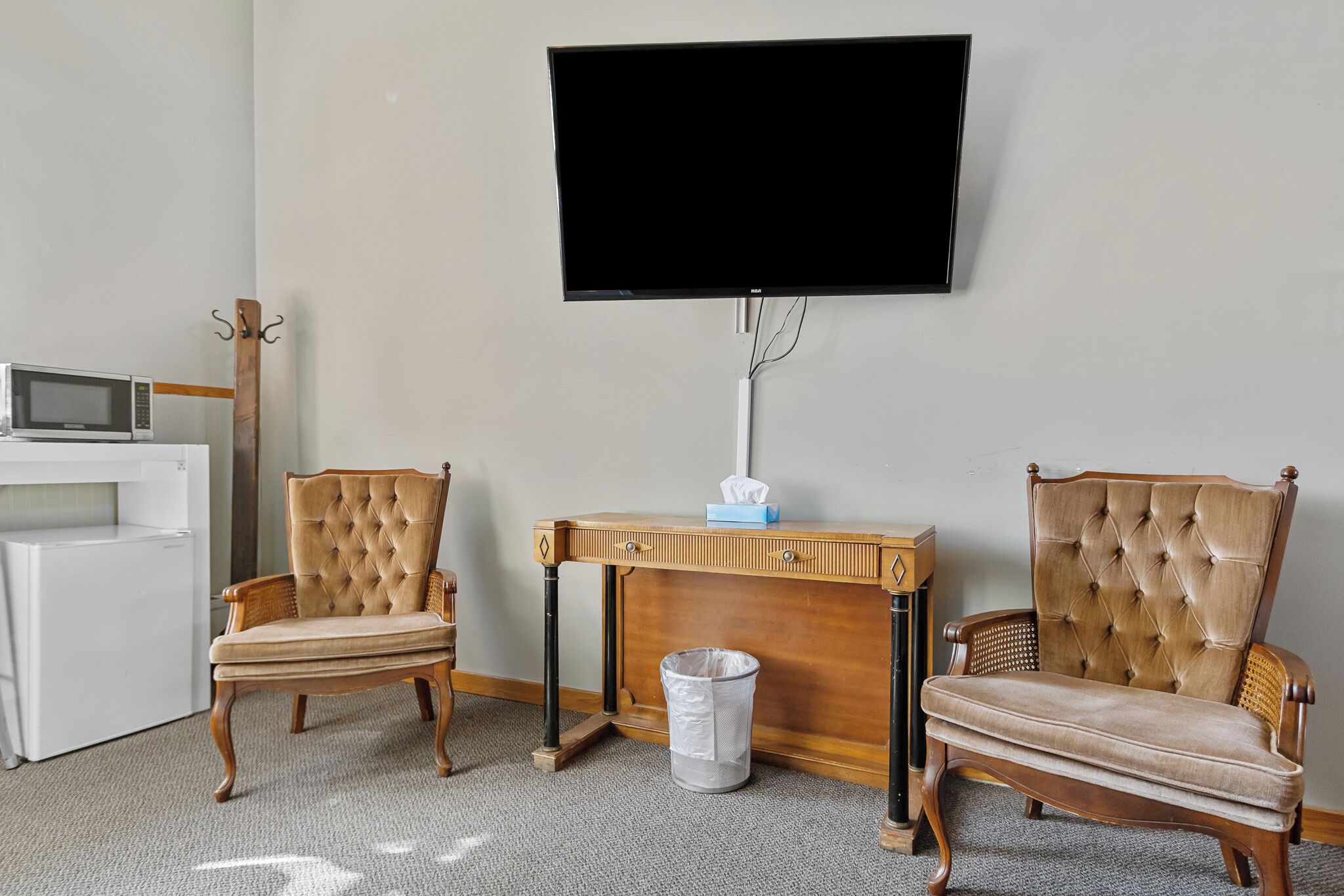 Une pièce équipée d’un téléviseur à écran plat fixé au mur, d’une console en bois sur laquelle se trouve une boîte de mouchoirs, de deux fauteuils rembourrés, d’un micro-ondes blanc et d’une poubelle.