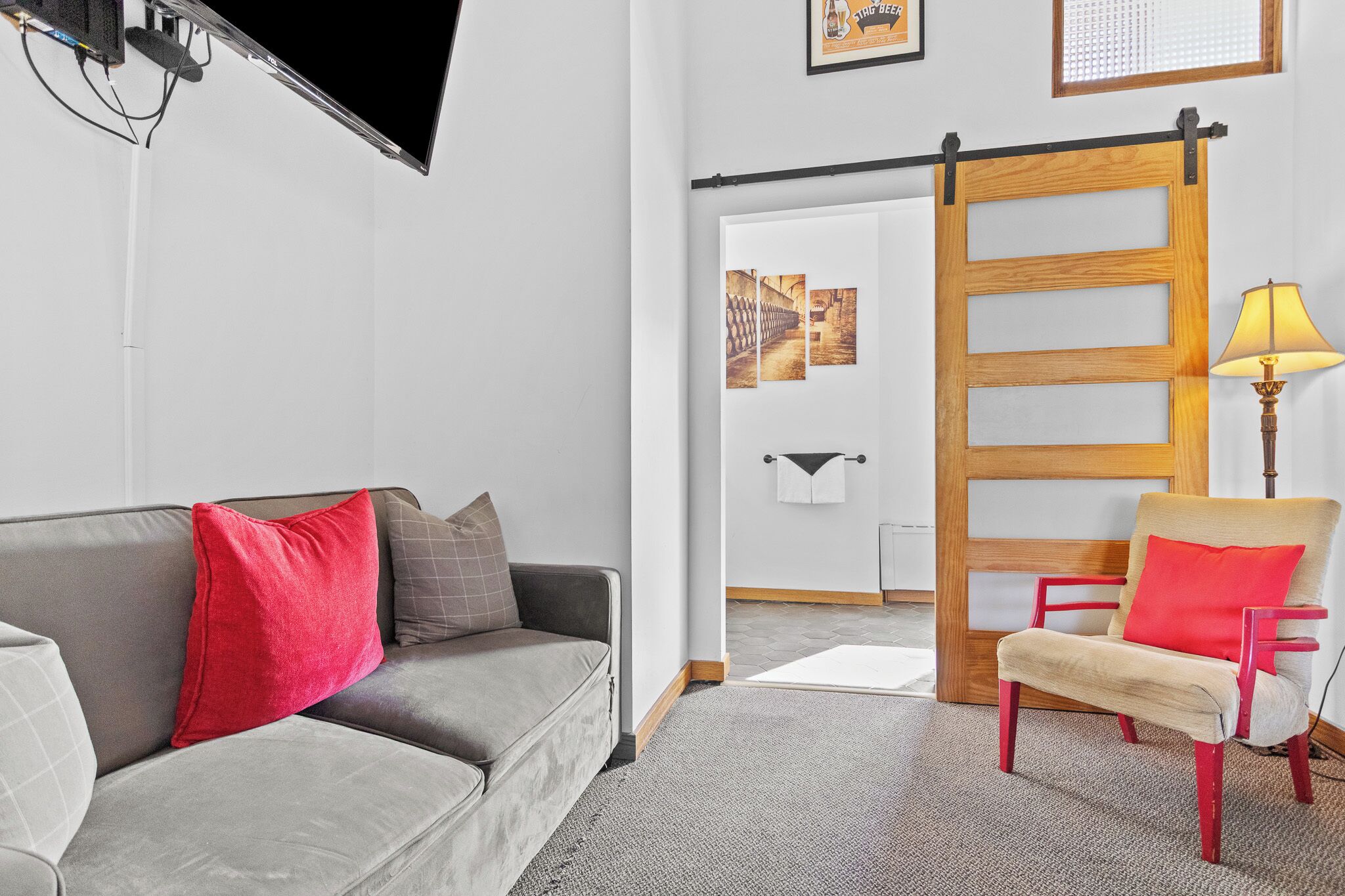 Un salon moderne avec un canapé gris, des coussins rouges et gris, un fauteuil rouge et beige, une porte coulissante en bois et une lampe.